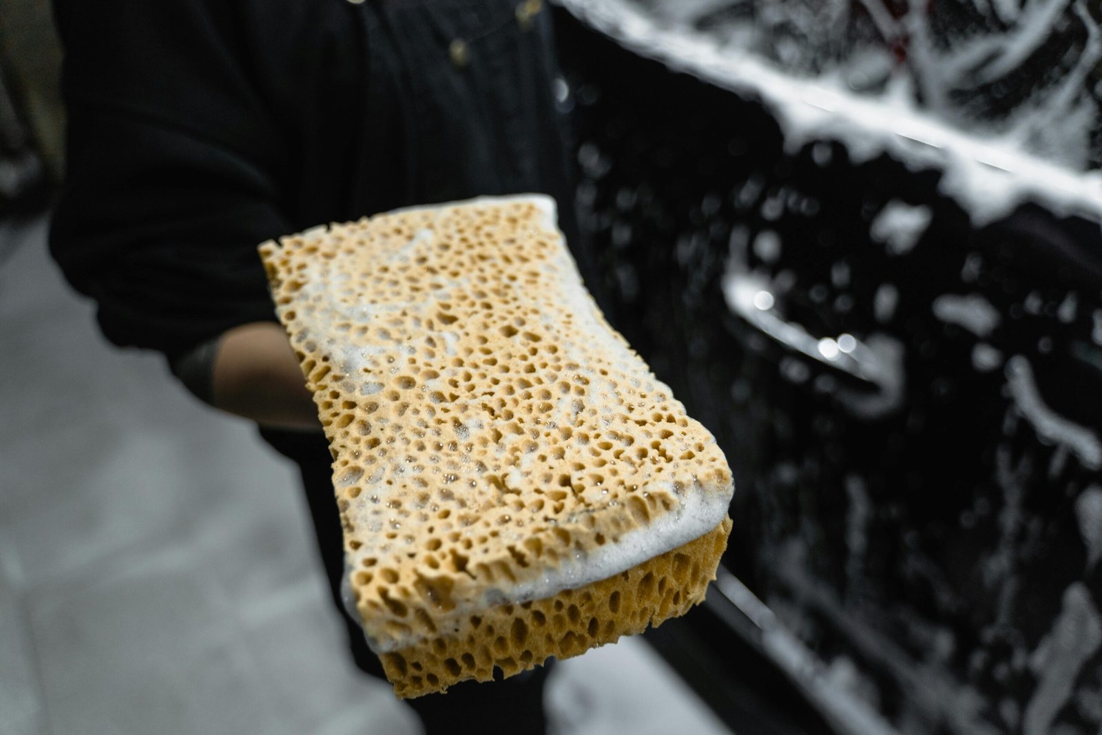 Sponge covered in soap suds held beside a car, highlighting car cleaning.